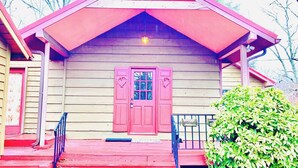 Un edificio de madera con puerta roja, un pequeño porche y un arbusto verde.