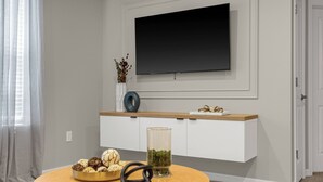 Uma sala de estar moderna com uma televisão de tela plana na parede, uma mesa de centro de madeira com objetos decorativos e um console branco com um vaso de flores secas.