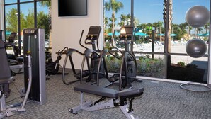 Uma academia com máquinas de cardio, um banco de musculação e uma grande janela com vista para uma piscina e palmeiras.
