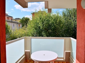 Ein Balkon mit Glastrennwand, einem runden Tisch und Blick auf Grünflächen und Gebäude.