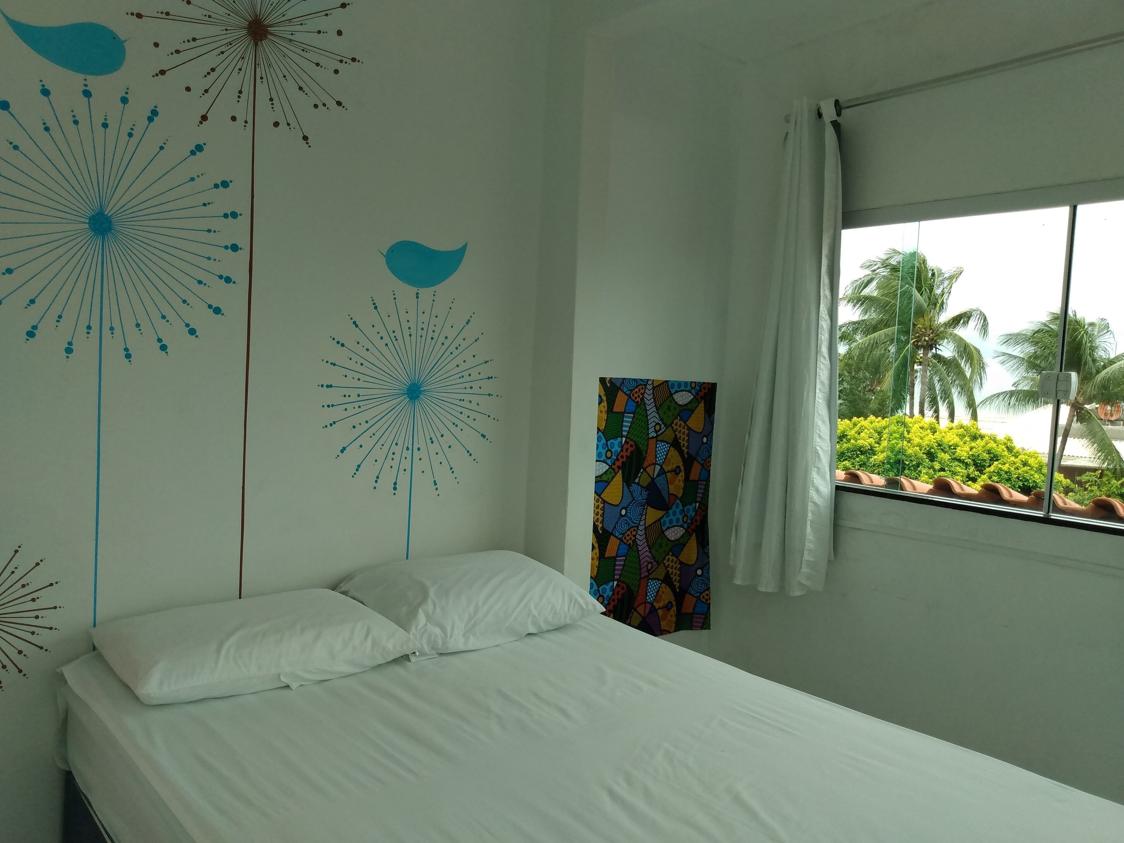 Een slaapkamer met een bed, witte kussens, een kleurrijk wandtapijt en een raam met uitzicht op palmbomen.