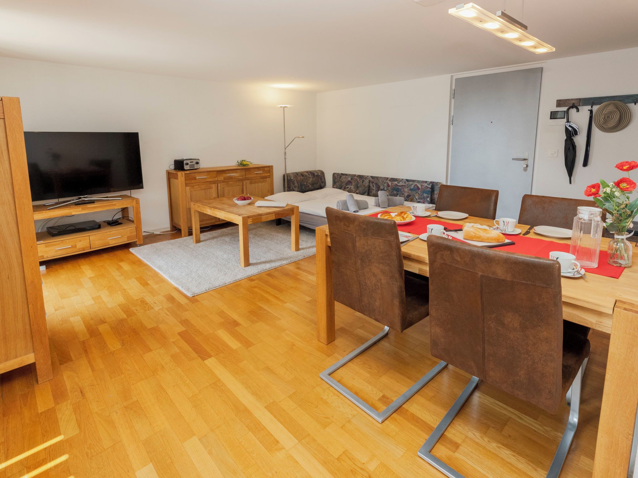 Ein modernes Wohnzimmer mit Flachbildfernseher, Holzmöbeln, einem Esstisch mit Stühlen und einer Couch.