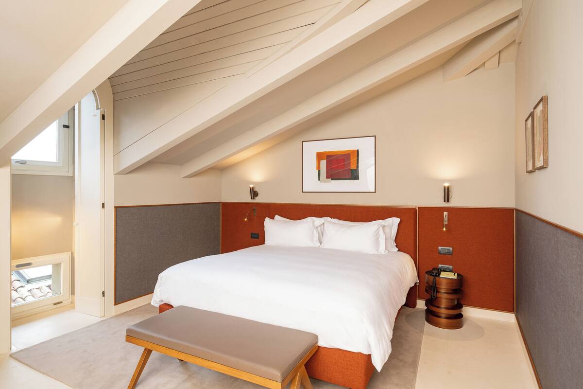 A modern hotel room with a large bed, a bench, a bedside table, and a framed artwork on the wall.