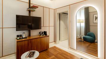 Una moderna camera d'albergo con una scrivania in legno, una TV a schermo piatto, un piccolo tavolino rotondo e uno specchio che riflette un'accogliente zona salotto con una lampada.