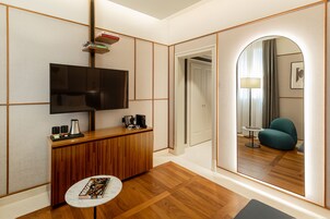 Una moderna camera d'albergo con una scrivania in legno, una TV a schermo piatto, un piccolo tavolino rotondo e uno specchio che riflette un'accogliente zona salotto con una lampada.