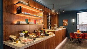 A well-lit bar area with wooden shelves displaying books and bottles, a buffet counter with various food items, and a seating area with chairs. - Hotel Brun (Bologna)