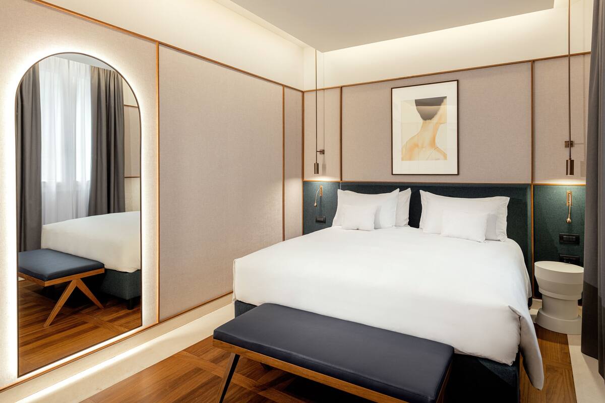 A modern hotel room with a large bed, bedside tables, a bench, a mirror, and a framed artwork on the wall.