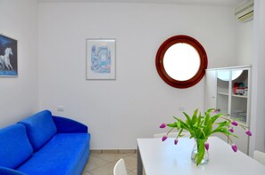 Una habitación con un sofá azul, una mesa blanca con un jarrón de flores, un espejo redondo y un armario con estantes.