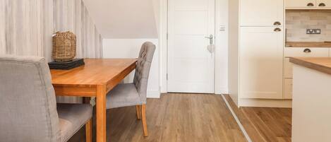 Una cocina compacta con armarios blancos, una mesa de comedor de madera y una silla tapizada de color gris.