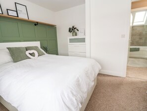 A bedroom with a bed, a green headboard, a bedside table with a vase, and a bathroom visible through an open door.