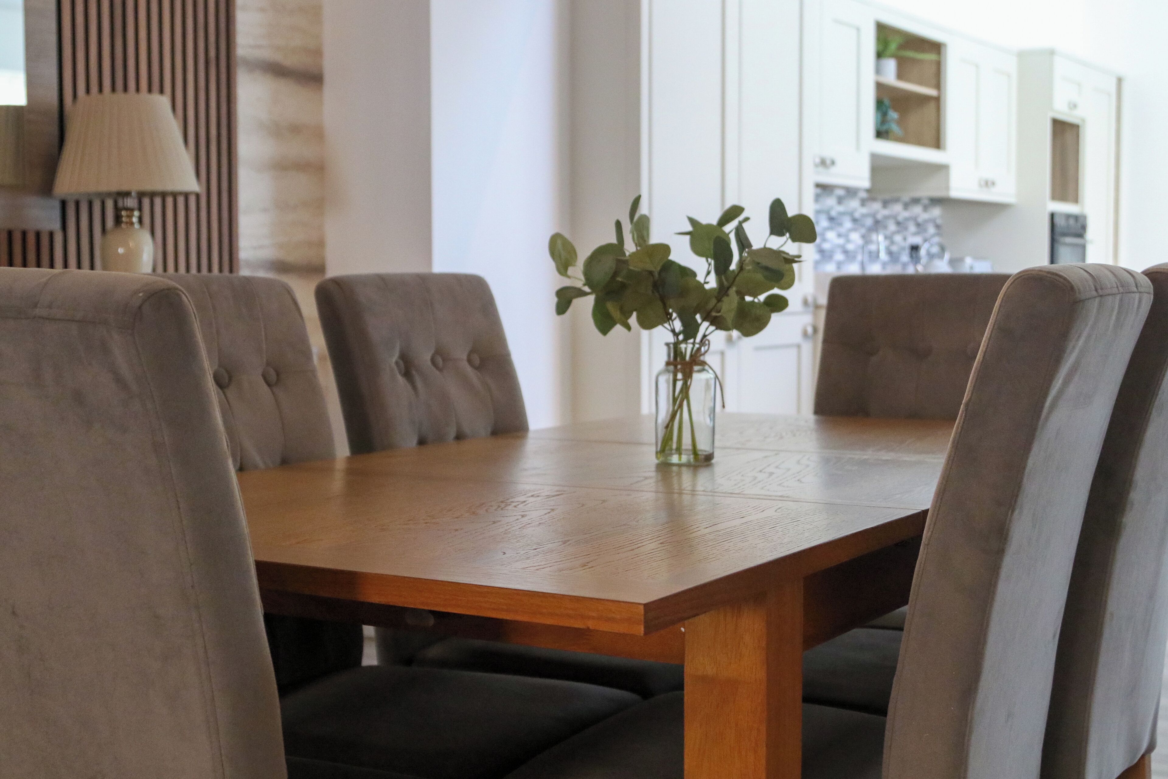 A dining table with four chairs, a vase with green leaves, and a lamp in the background.