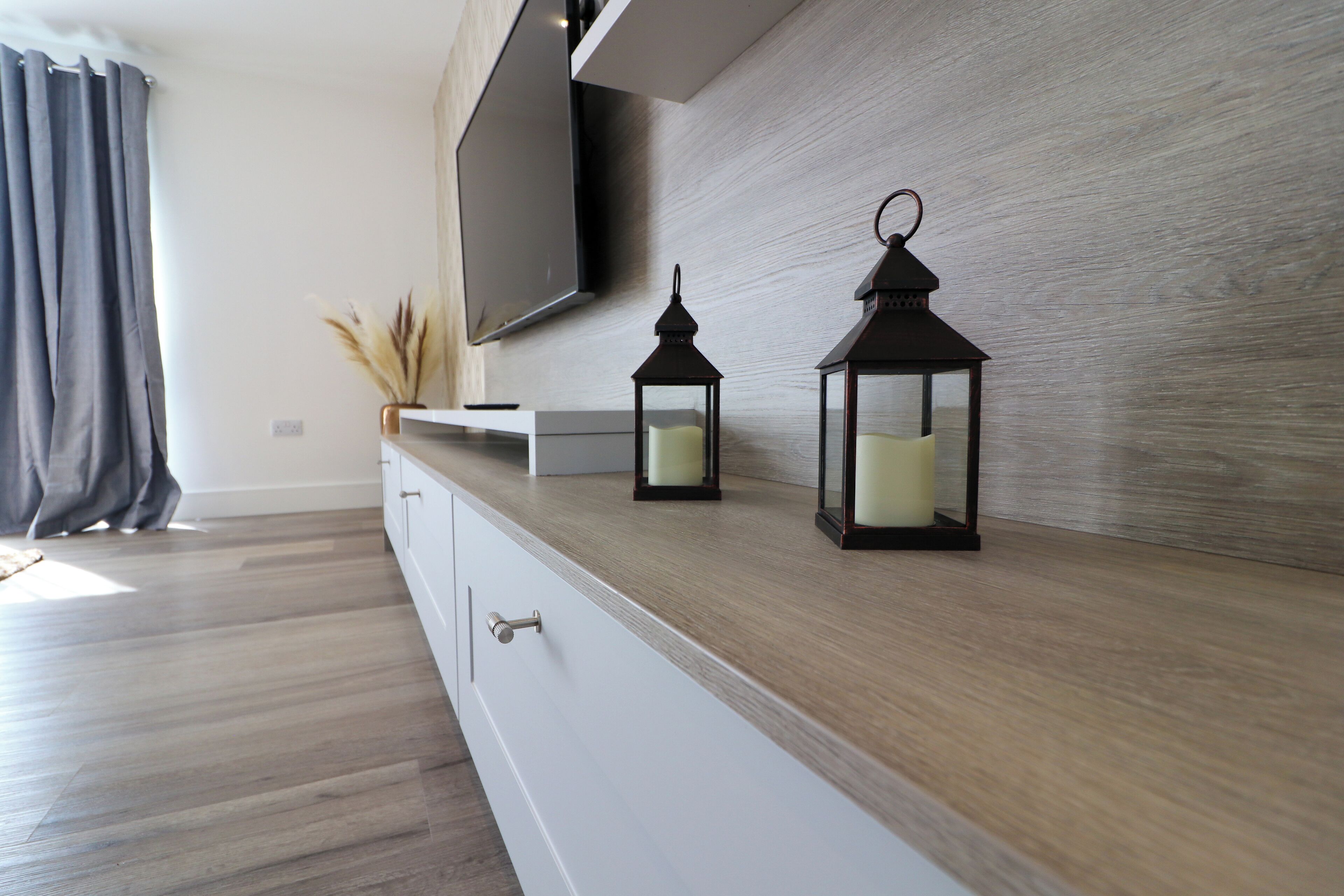 Een moderne woonkamer met houten vloer, een flatscreen televisie aan de muur en twee zwarte lantaarns op een houten console tafel.