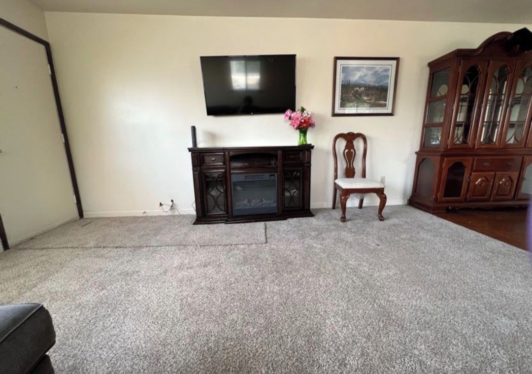 A living room with a flat-screen TV mounted on the wall, a wooden cabinet, a chair, and a carpeted floor.