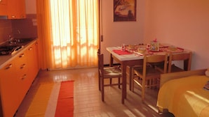 Una cocina compacta con estufa, pileta y armarios color naranja. Hay un comedor con una mesa dispuesta para dos personas, una silla y una cama con colcha amarilla.