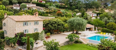 Une maison en pierre avec une piscine, entourée d’une végétation luxuriante et d’autres maisons sur une colline.