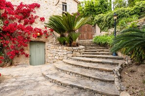 Un escalier en pierre mène à un bâtiment en pierre doté d’une porte verte, le tout étant entouré d’une végétation luxuriante et de fleurs rouges.