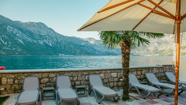 A terrace with lounge chairs and a palm tree, overlooking a lake with mountains in the background.
