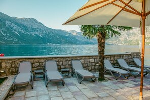 Une terrasse avec des fauteuils et un palmier, donnant sur un lac avec des montagnes en arrière-plan.