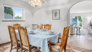 Comedor con una mesa redonda dispuesta para seis personas, rodeada de sillas de madera con asientos de mimbre. Un candelabro cuelga del techo, y hay una planta enmarcada en la pared.