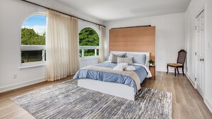 Un dormitorio con una cama grande, dos ventanas con cortinas, un cabecero de madera, una silla y vistas al verde exterior.