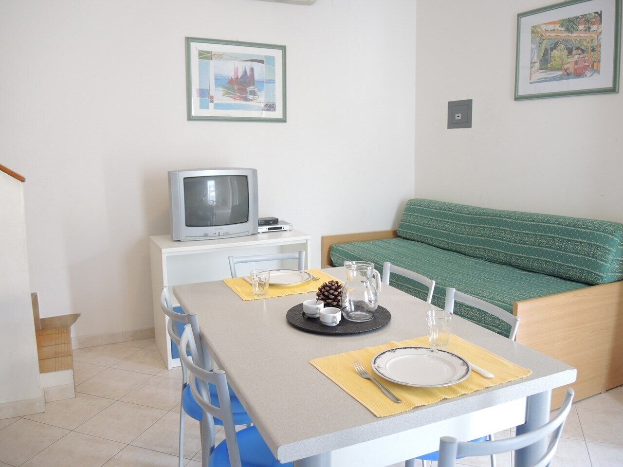A dining area with a table set for two, a television on a stand, and a green sofa in the background.