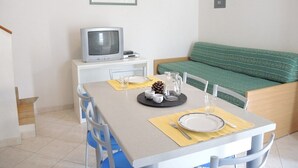 Ein Essbereich mit einem für zwei Personen gedeckten Tisch, einem Fernseher auf einem Schrank und einem grünen Sofa im Hintergrund.