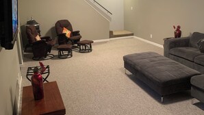 Una sala de estar con un sofá gris, un reposapiés gris, un sillón marrón, una mesita con un jarrón y una escalera que lleva a otro nivel.