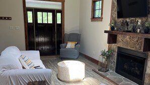 A spacious living room with a stone fireplace, a large flat-screen TV mounted on the wall, and a comfortable seating area with two sofas and a chair.