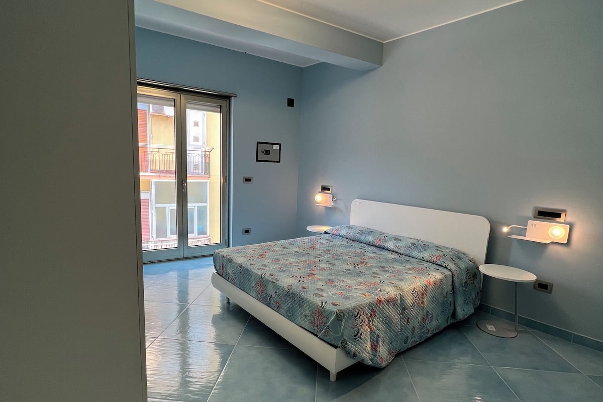 A bedroom with a bed, a nightstand, a wall-mounted light, and a door leading to a balcony.