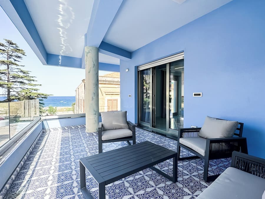 Uma varanda com chão azul e branco estampado, duas cadeiras, uma mesa e vista para o mar.
