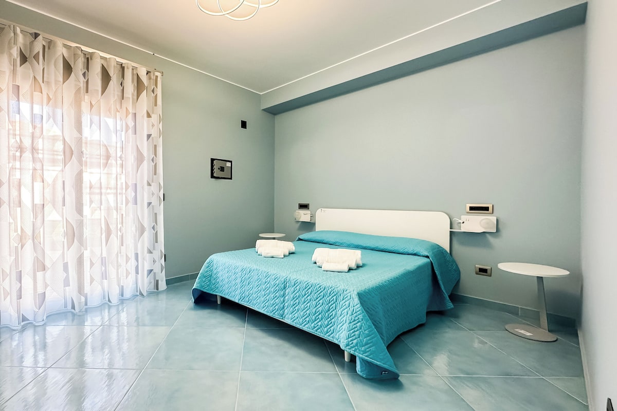 A bedroom with a bed, a bedside table, a nightstand, and a window with curtains.