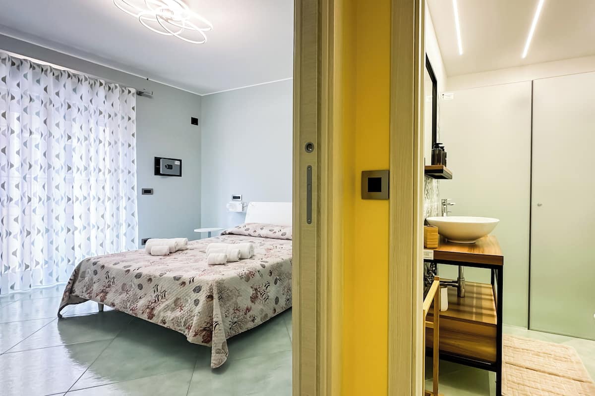 A hotel room with a bed, a bedside table with a sink, a toilet, and a shower area.