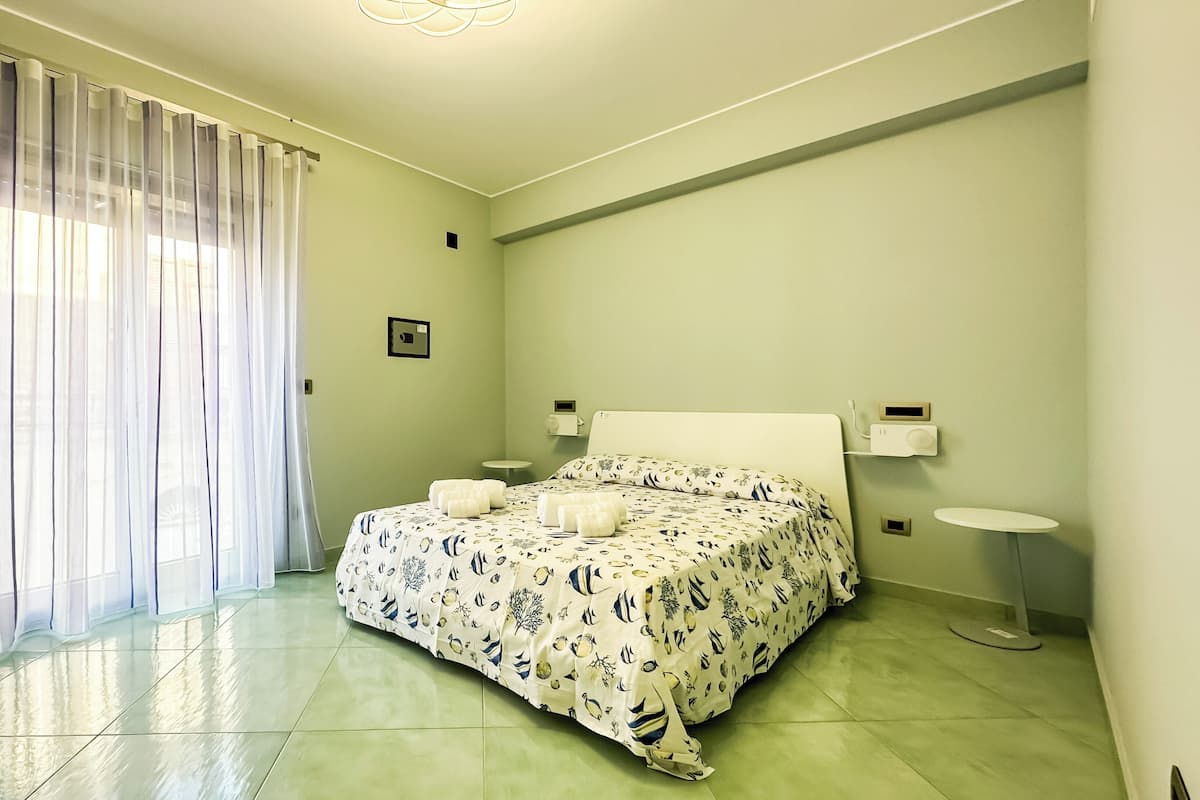 A bedroom with a bed, two bedside tables, a nightstand, a window with sheer curtains, and a light switch on the wall.