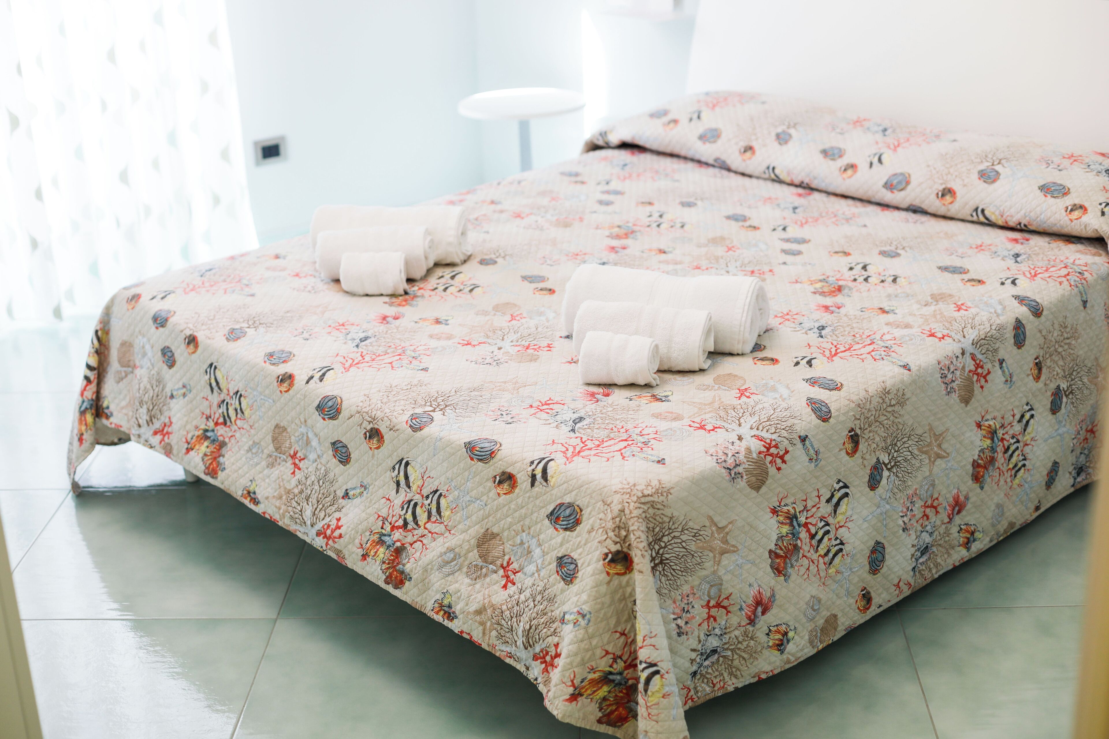 A neatly made bed with a floral and sea-themed bedspread, two rolled towels, and a white bedside table.