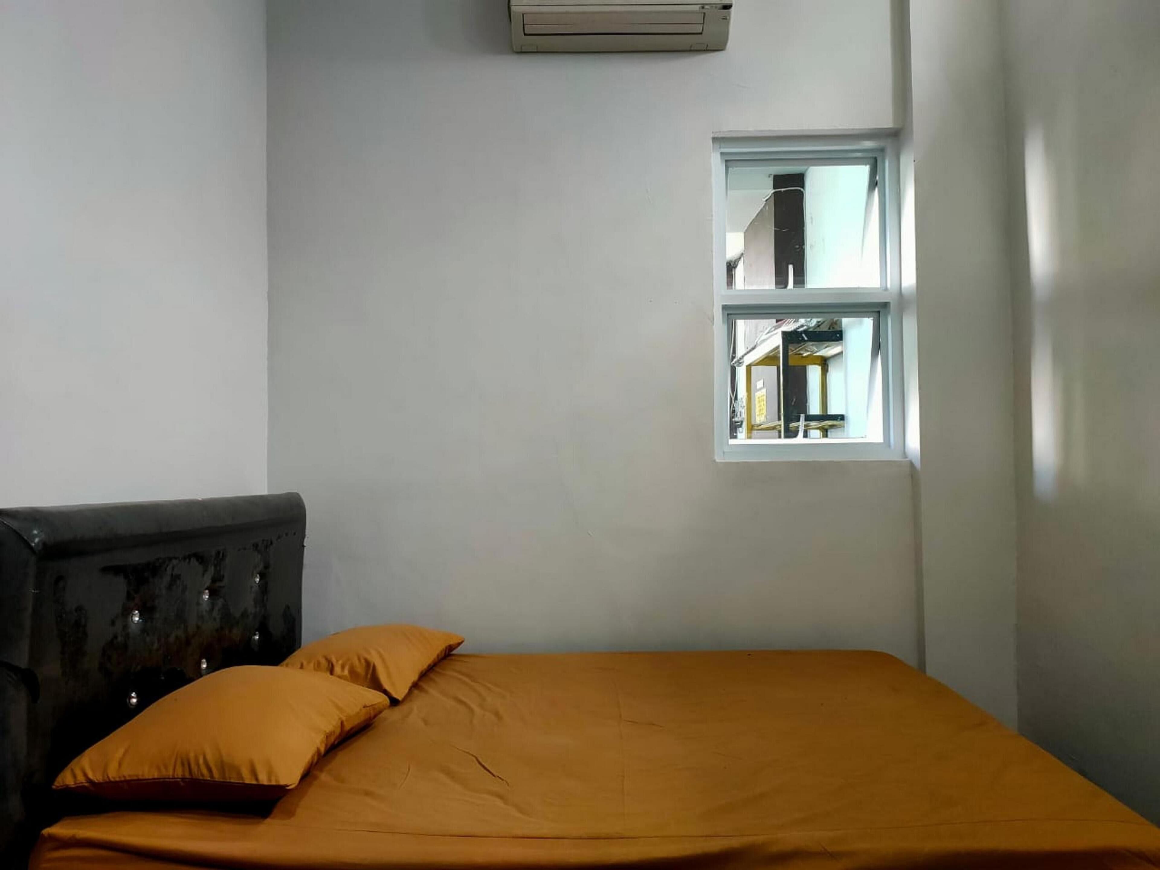 Un lit simple avec une couvre-lit orange et deux oreillers, une fenêtre donnant sur l’extérieur et un climatiseur fixé au mur.