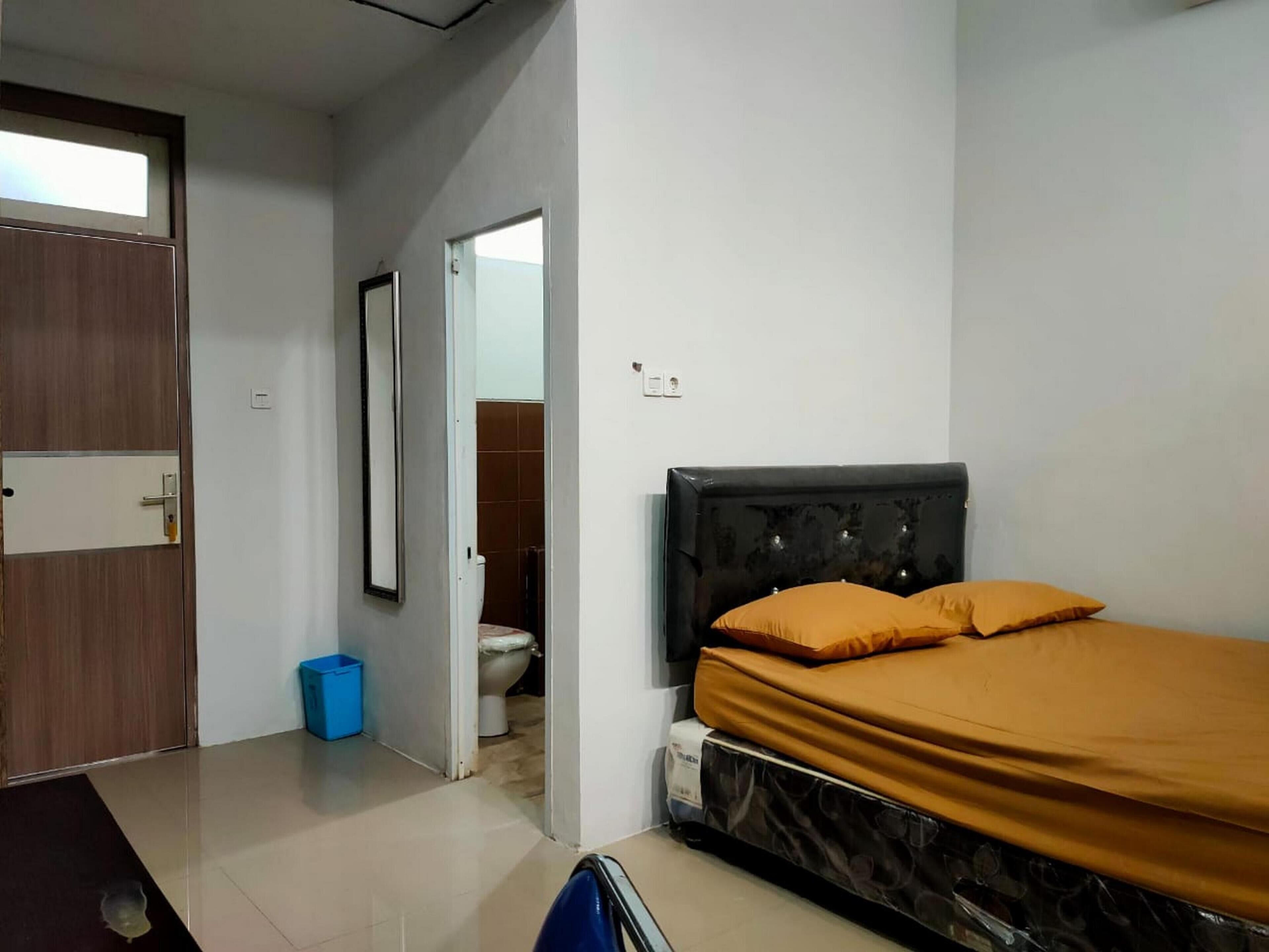 Ein Schlafzimmer mit einem Bett, einer Holztür, einem blauen Mülleimer und einer Toilette, die durch eine geöffnete Tür sichtbar ist.