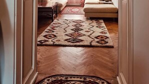 Un couloir mène à un salon où se trouvent un canapé, une petite table et une lampe. Un tapis à motifs recouvre le sol, et une fenêtre est agrémentée de rideaux.