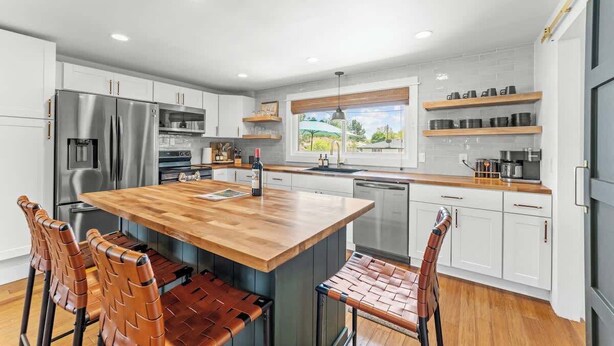 Une cuisine moderne avec des armoires blanches, des appareils en acier inoxydable, un îlot central en bois et une vue sur l’extérieur.