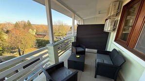 A balcony with wicker furniture, a large window, and a view of trees and buildings. - Holiday apartment with outdoor pool and air conditioning (Balatonföldvár)