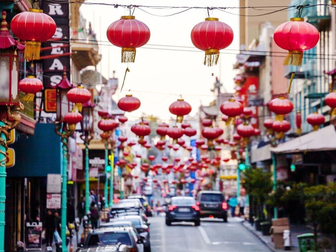 A street lined with red lanterns, cars parked on the side, and buildings with signs.