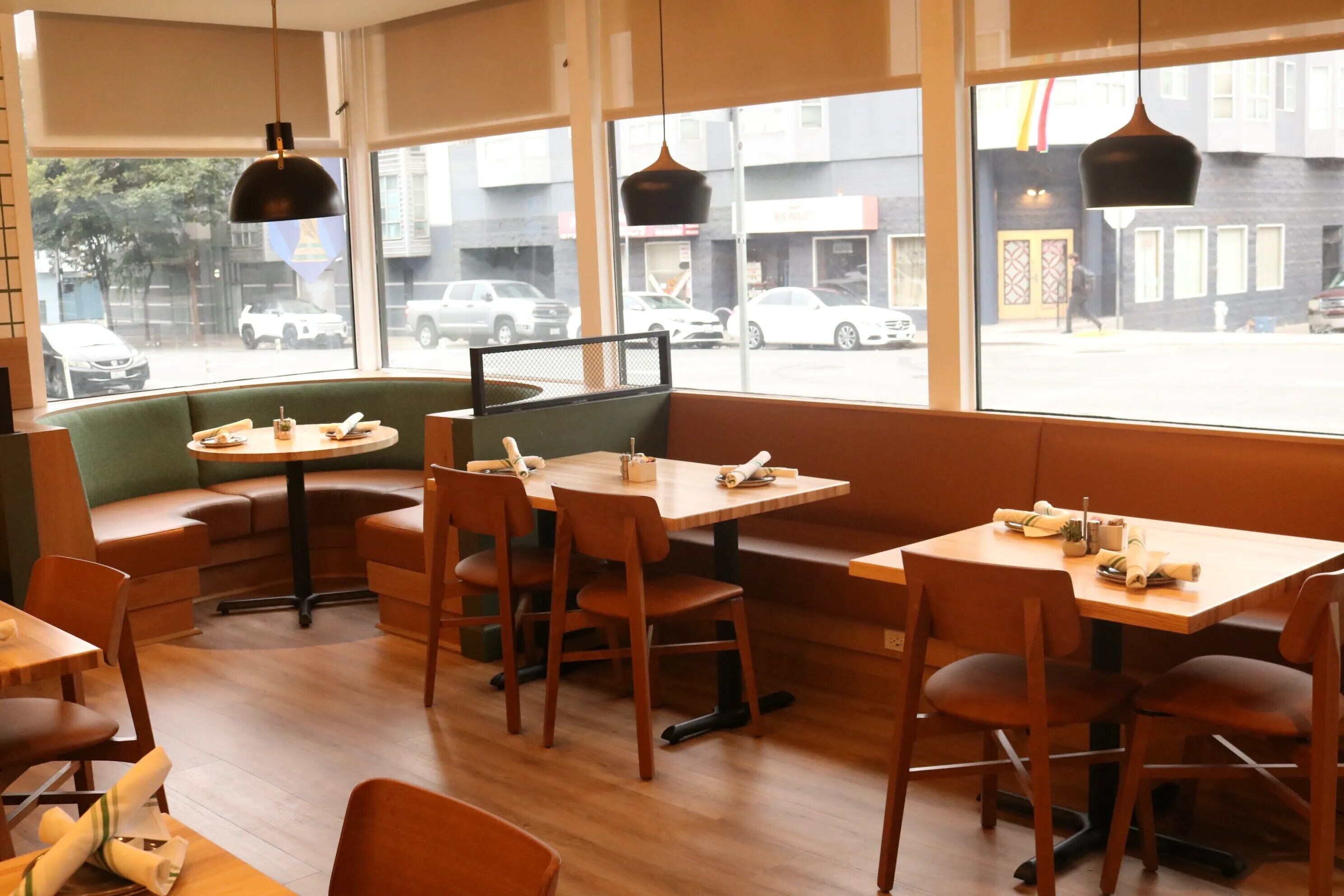Un espace de détente intérieur avec des tables et des chaises en bois, un banc rembourré de couleur verte et de grandes fenêtres donnant sur la rue.