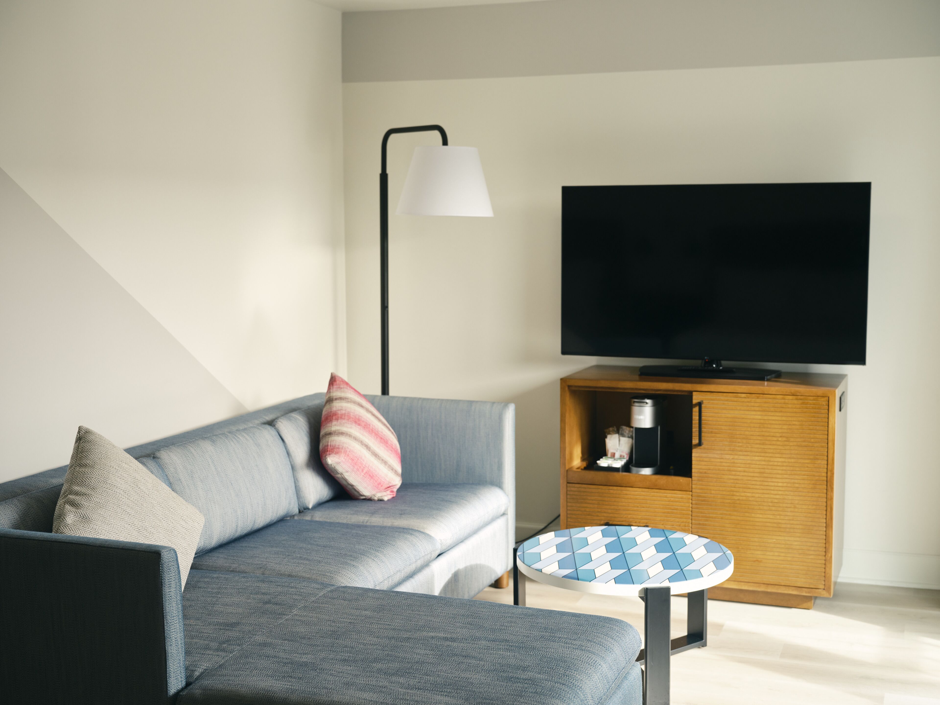 A modern living room with a grey sofa, a small round coffee table, a flat-screen TV on a wooden cabinet, and a floor lamp.