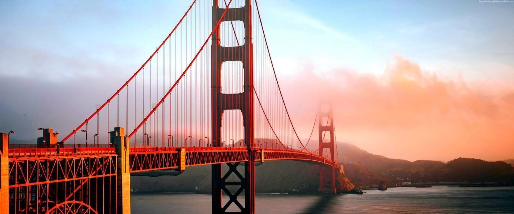 A suspension bridge with a central tower and cables, spanning over a body of water with hills in the background.