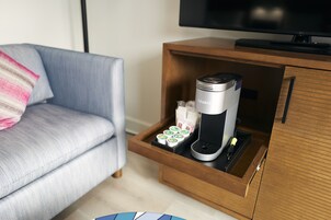 A compact coffee maker in a drawer with a remote control, a TV above it, and a sofa with a striped pillow in the background.