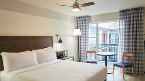 A hotel room with a large bed, a ceiling fan, a desk with a lamp, a chair, a small table, and a view of the outside balcony. - Soma House (San Francisco)