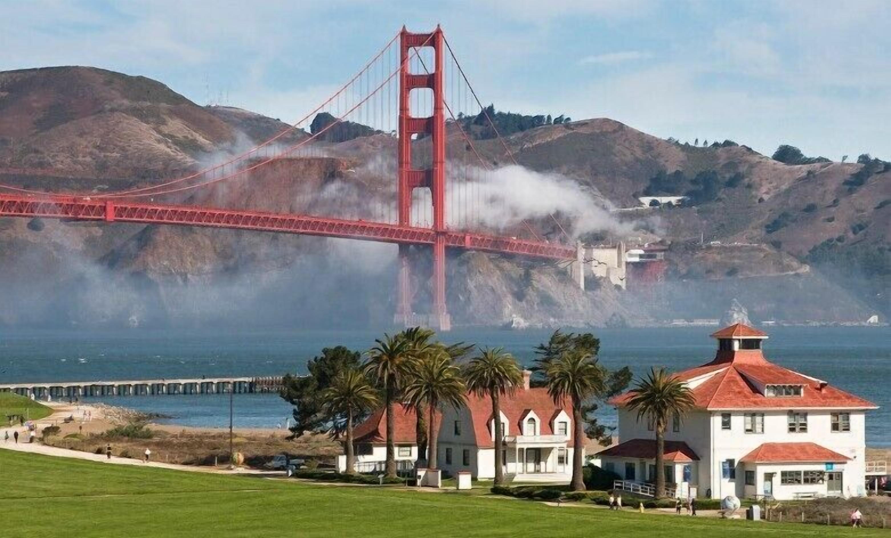 Vista da Ponte Golden Gate com vapor subindo da estrutura, sobre uma área costeira com palmeiras e um edifício branco com telhado vermelho.