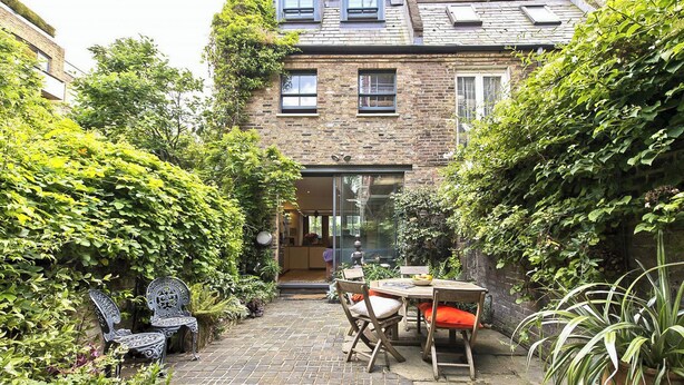 A brick house with a small patio area featuring a table and chairs, surrounded by lush greenery.