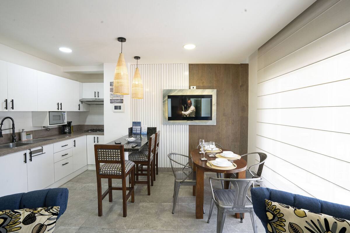 A modern kitchen with white cabinets, a dining area with a wooden table and chairs, and a television mounted on the wall.