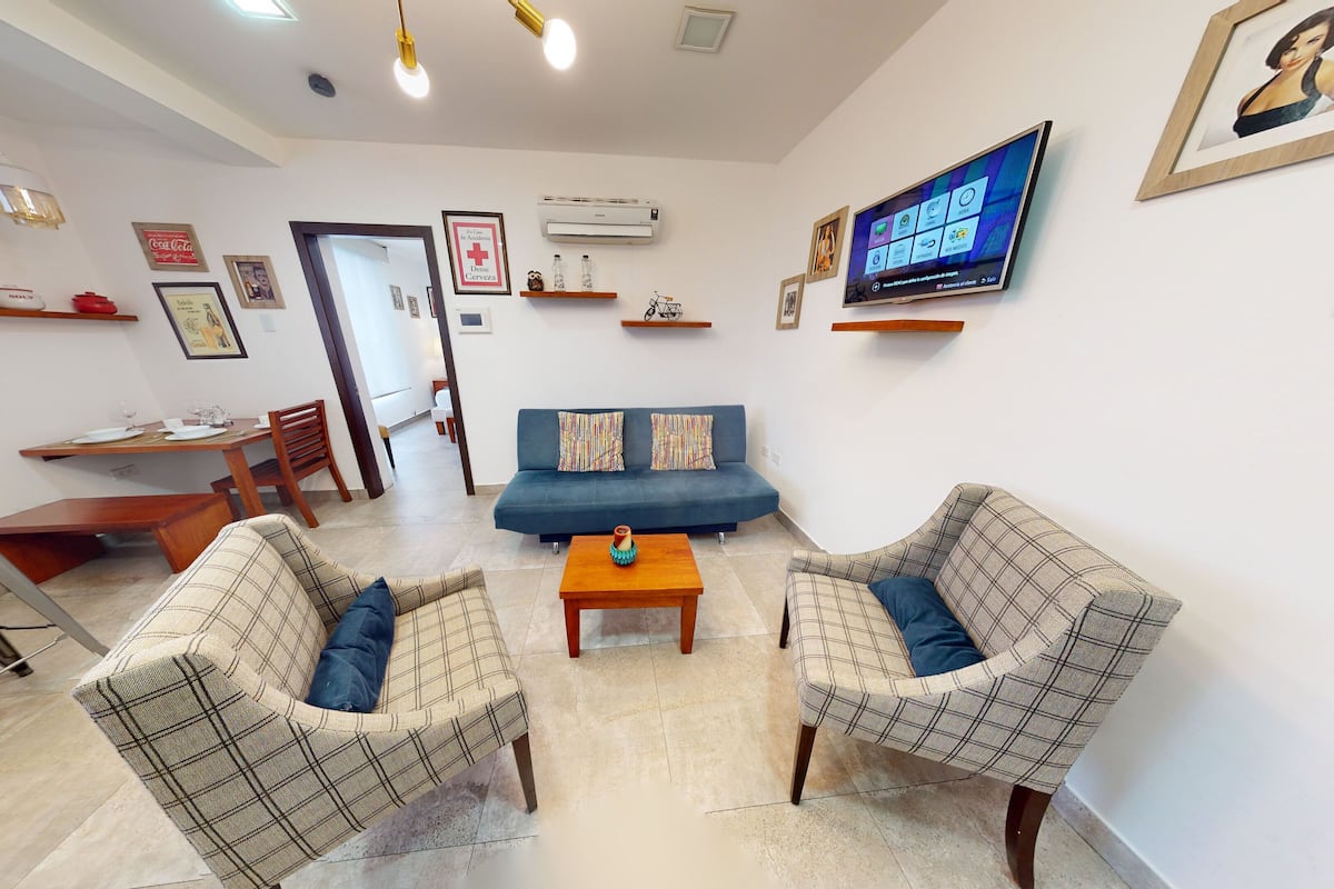 A living room with a sofa, two armchairs, a coffee table, and a TV. There is a dining area with a table and chairs, and a kitchen area with a stove and sink.