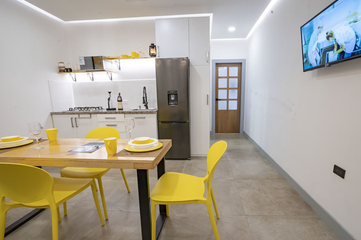A modern kitchen with a dining table set for four, yellow chairs, a stainless steel refrigerator, and a flat-screen TV mounted on the wall.